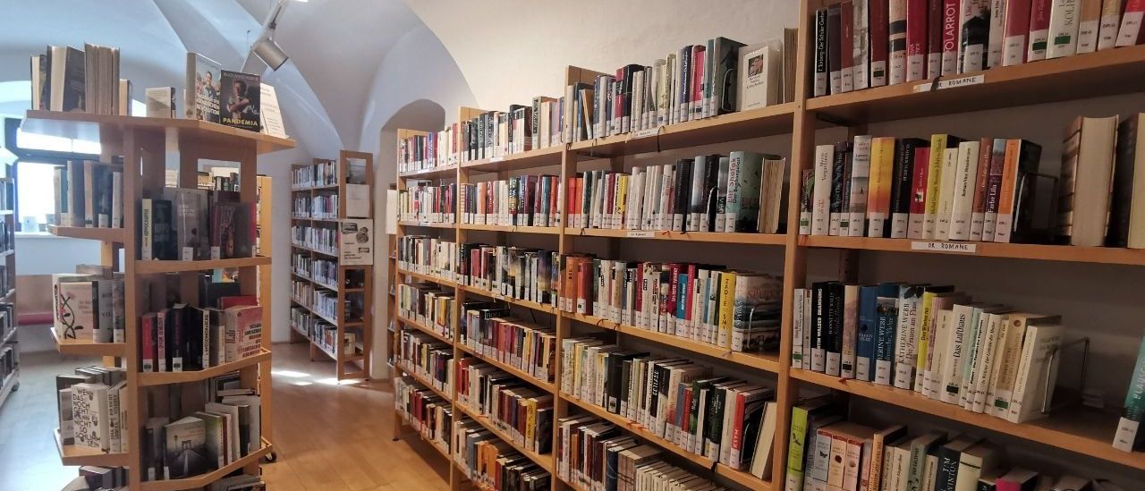 Eine Bibliothek mit holzfarbenen Regalen voller Bücher, einer Decke mit einer Lichtarmature und einem hölzernen Boden.