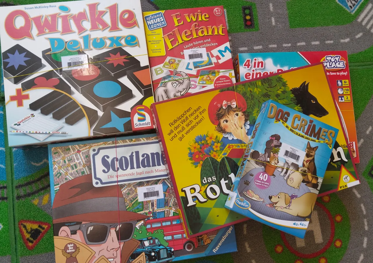 Eine Sammlung von Brettspielen mit bunten Illustrationen und deutschen Texten auf den Schachteln. Titel sind 'Ewie Elefant', 'Scotland' und 'Dog Crime'. Die Spiele sind auf einem grünen Teppich platziert.