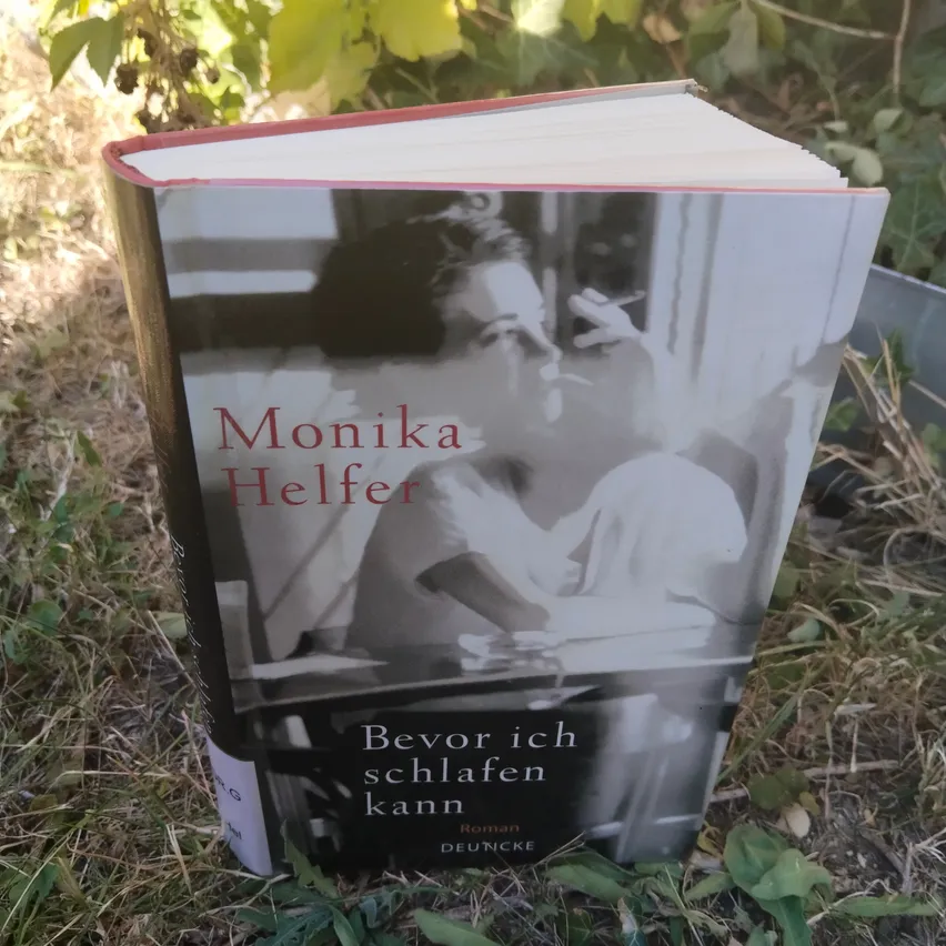 Ein Buch mit dem Titel 'Bevor ich schlafen kann' von Monika Helfer, das ein Schwarz-Weiß-Bild einer rauchenden Frau zeigt, liegt auf dem Gras.