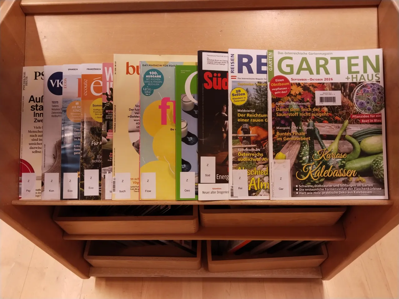 Ein Holzregal zeigt eine Reihe von Zeitschriften, darunter Titel wie 'GARTEN', 'REISEN' und 'SUD', mit Etiketten, die ihre Kategorien wie 'Z', 'Eco' und 'Flow' angeben.