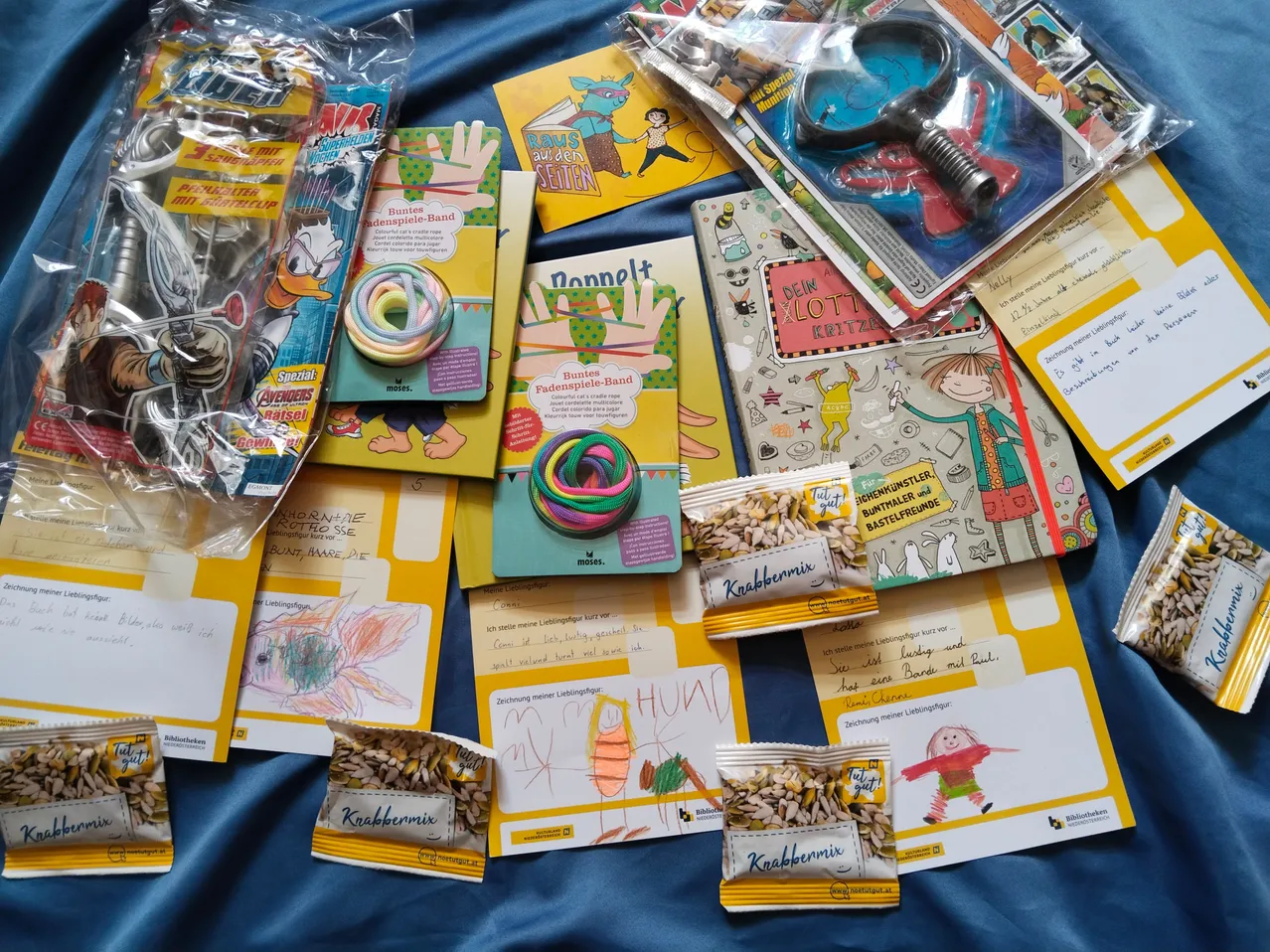 Eine Sammlung von bunten Kinderbüchern, Spielzeug und Snacks auf einer blauen Oberfläche. Die Artikel umfassen eine Lupe, ein Buch mit dem Titel 'RAWS auf den Seiten' und einen Beutel mit gemischten Snacks.