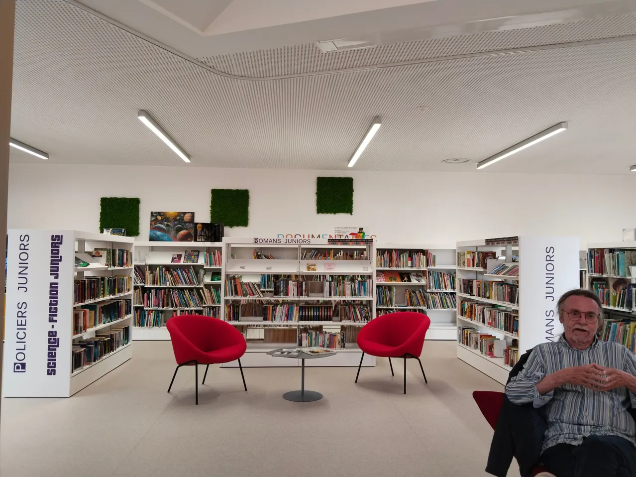 Eine Innenbibliothek mit Bücherregalen, roten Stühlen, einem Tisch und einem sitzenden Mann.