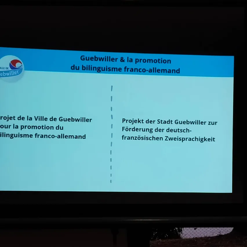 Ein Beamer zeigt eine Folie mit einem Titel auf Französisch und Deutsch, 'Guebwiller & la promotion du bilinguisme franco-allemand'. Unter dem Titel befinden sich zwei Textabschnitte, einer auf Französisch und der andere auf Deutsch.