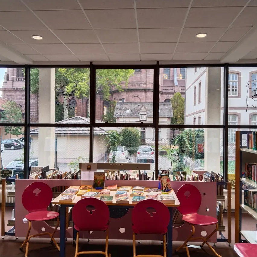 Ein Innenansicht einer Bibliothek mit großen Fenstern, die einen Tisch mit Büchern, rote Stühle und Bücherregale voller Bücher zeigt. Draußen sind geparkte Autos und ein Gebäude mit einer Ziegelfassade sichtbar.