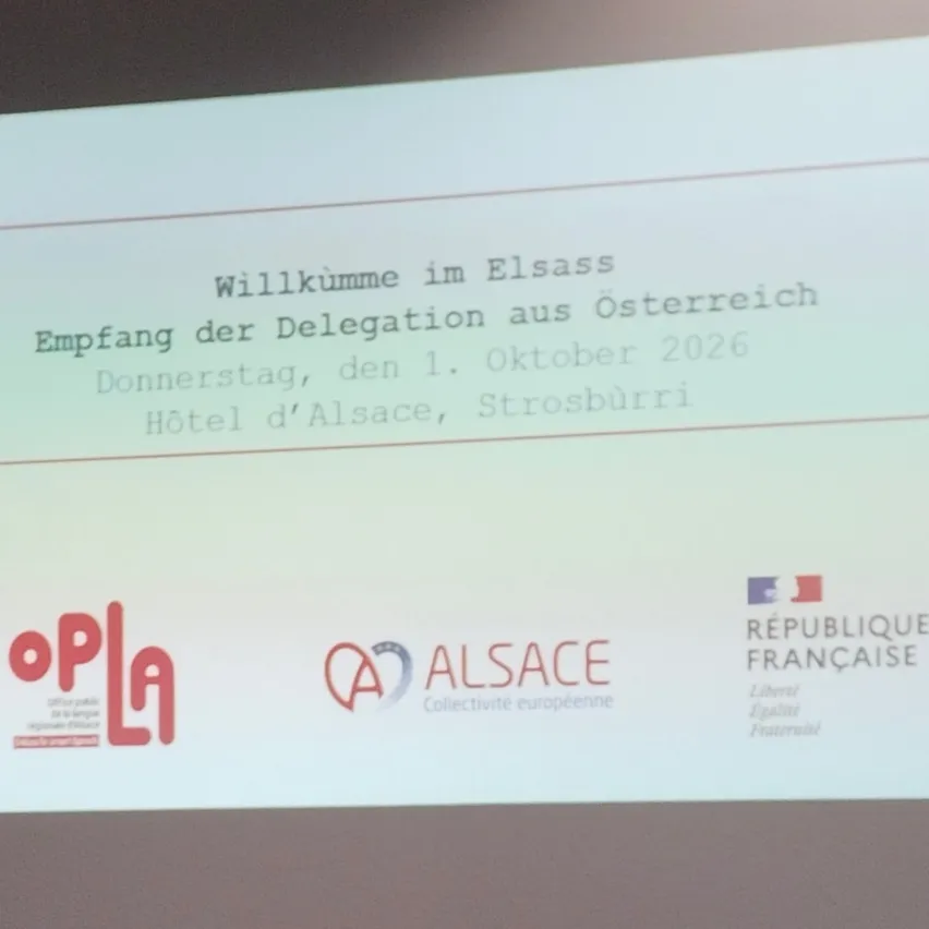 Eine Folie zeigt eine Willkommensnachricht für eine Delegation aus Österreich, datiert auf den 1. Oktober 2026. Der Ort ist Hotel d'Alsace, Straßburg. Logos für OPla, ALSA und REPUBLIQUE FRANCAISE sind vorhanden.