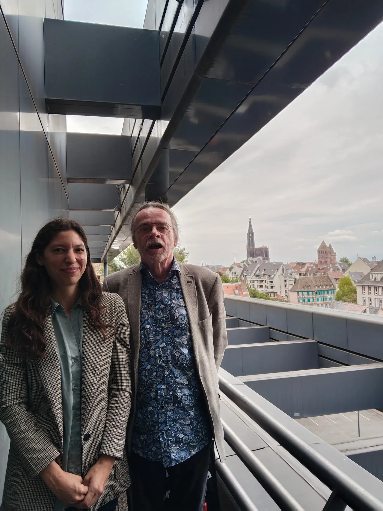 Ein Mann und eine Frau stehen auf einem Balkon, lächeln und schauen in die Kamera. Hinter ihnen ist ein Blick auf eine Stadt mit Gebäuden und einem bewölkten Himmel.