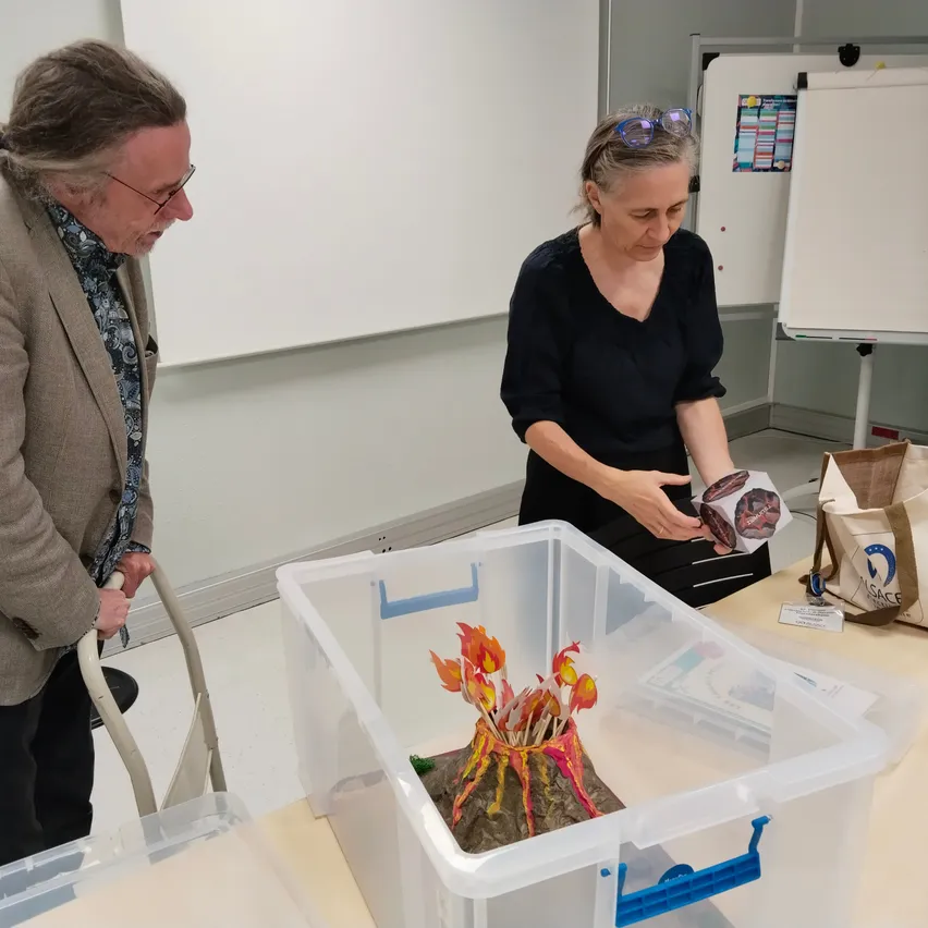 Ein Mann und eine Frau stehen in einem Raum. Der Mann schaut auf ein Vulkanmodell in einer Plastikbox auf einem Tisch. Die Frau hält ein Stück Papier und schaut auf den Vulkan.