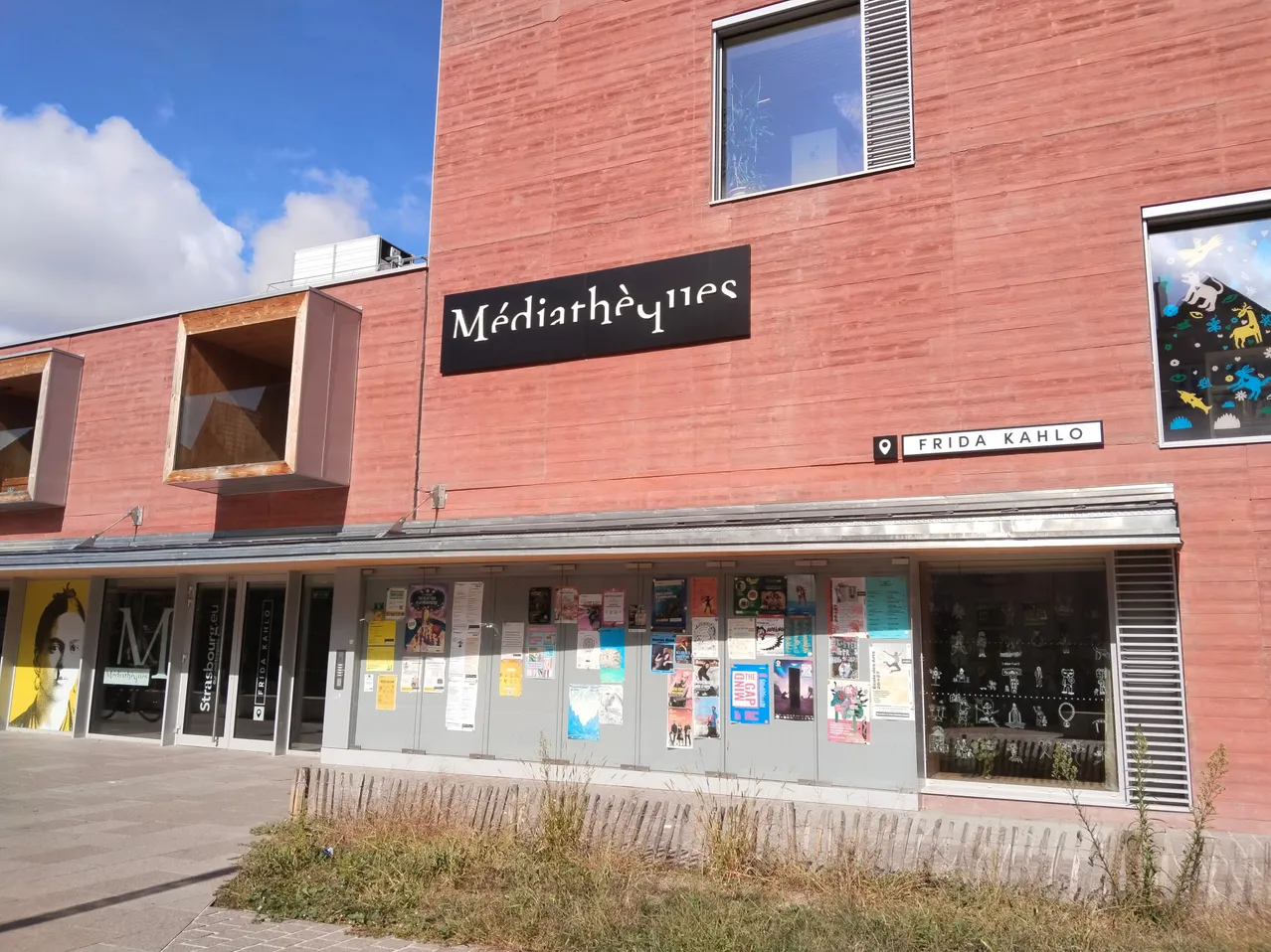 Ein rotes Gebäude mit einem Schild, das Médiathèques steht. Das Gebäude hat Glasfenster und Türen und ein Straßenschild, das Frida Kahlo steht.