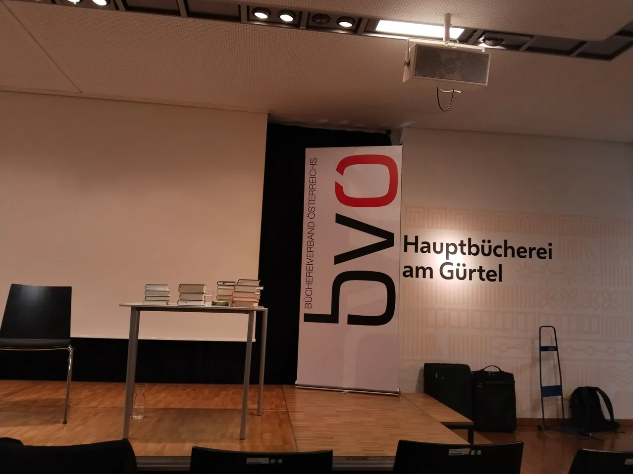Eine leere Bühne mit einem Tisch, auf dem Bücher stehen, und einem Banner mit dem Text '5V0' und 'Hauptbücherei am Gürtel'.