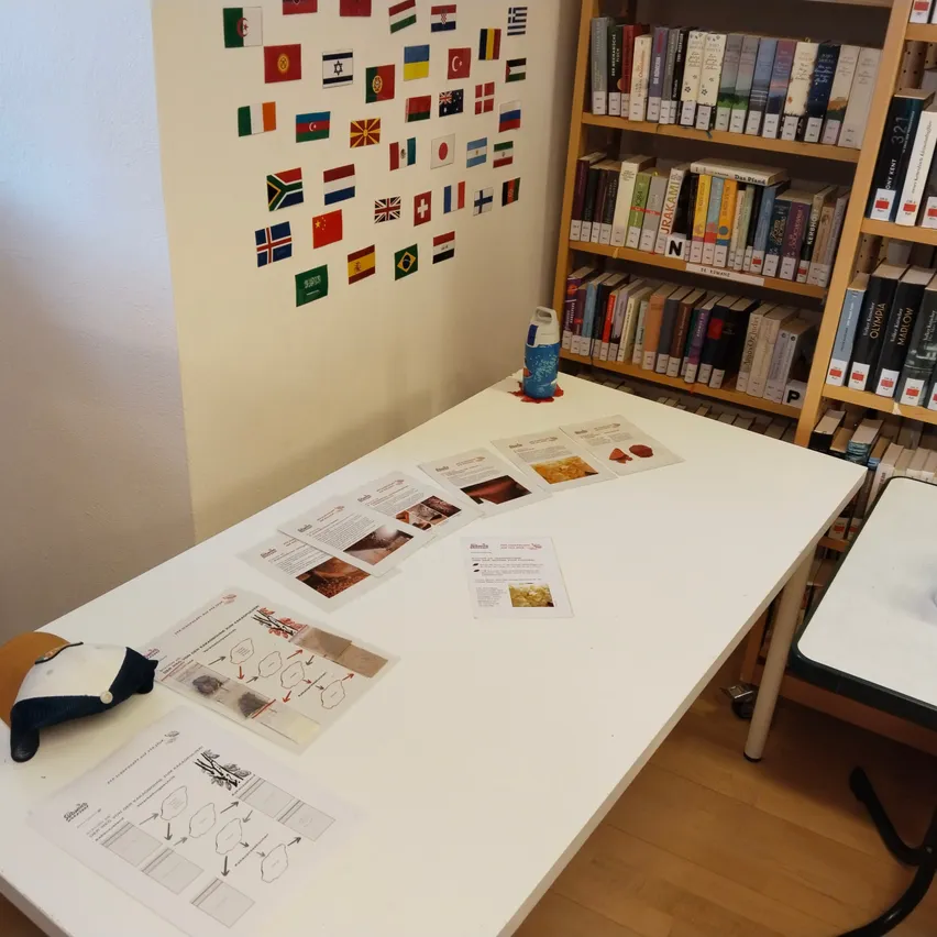 Ein weißer Tisch mit verschiedenen Papieren und einem blauen Behälter darauf. Hinter dem Tisch befindet sich eine Wand mit Flaggen verschiedener Länder. Auf der rechten Seite befindet sich ein Holzregal mit Büchern.