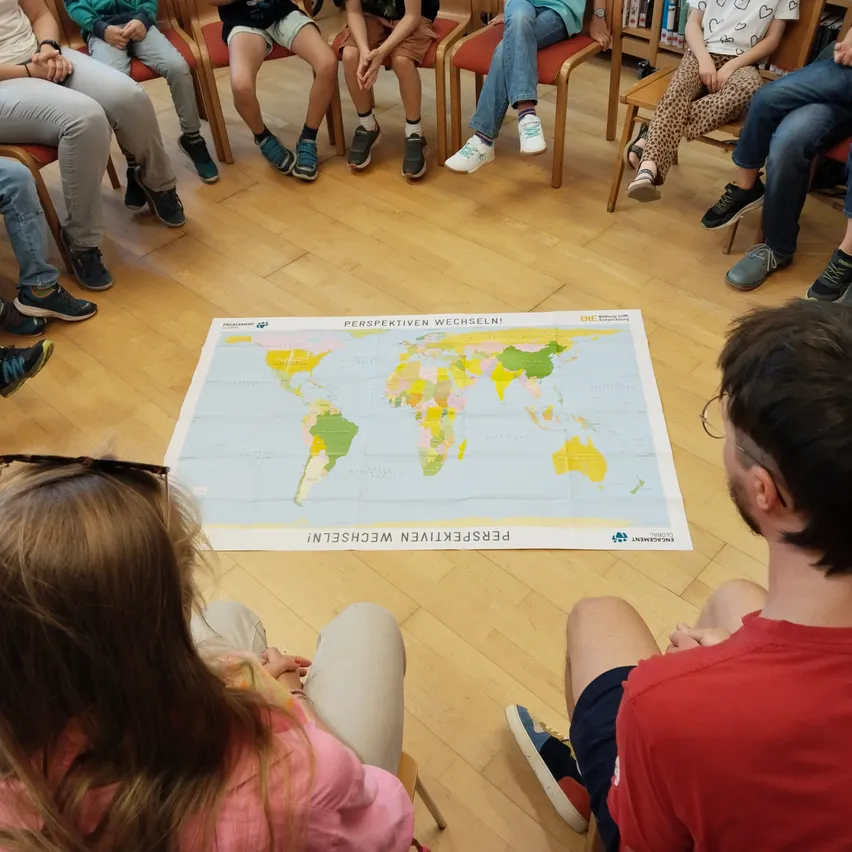 Eine Gruppe von Menschen sitzt im Kreis um eine große Weltkarte auf dem Boden.