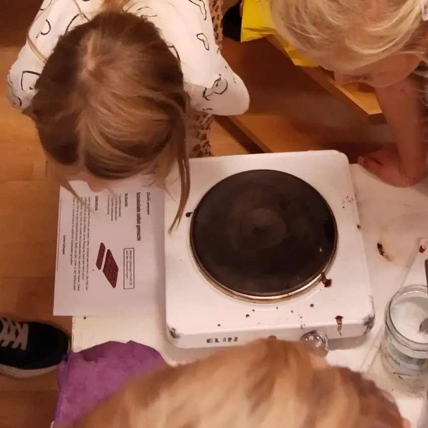 Eine Gruppe von Kindern versammelt sich um einen kleinen Elektroherd, wobei ein Mädchen einen Rezeptzettel genau betrachtet.