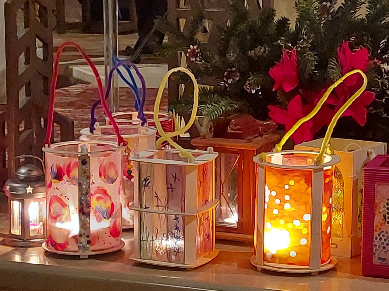 Mehrere dekorative Laternen mit bunten Malereien und Henkeln sind auf einem Tisch beleuchtet. Dahinter steht ein Weihnachtsbaum mit roten Blumen, und eine Person ist teilweise zu sehen.