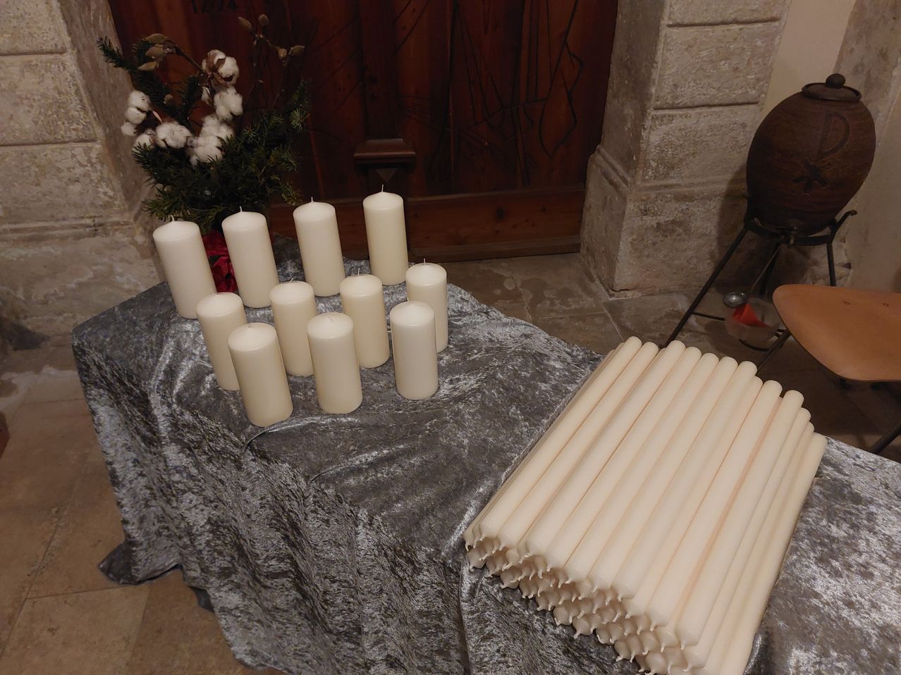 Ein Tisch mit einer silbernen Tischdecke ist mit mehreren weißen Kerzen bestückt, einige davon angezündet, andere nicht, mit einem Blumenarrangement im Hintergrund.