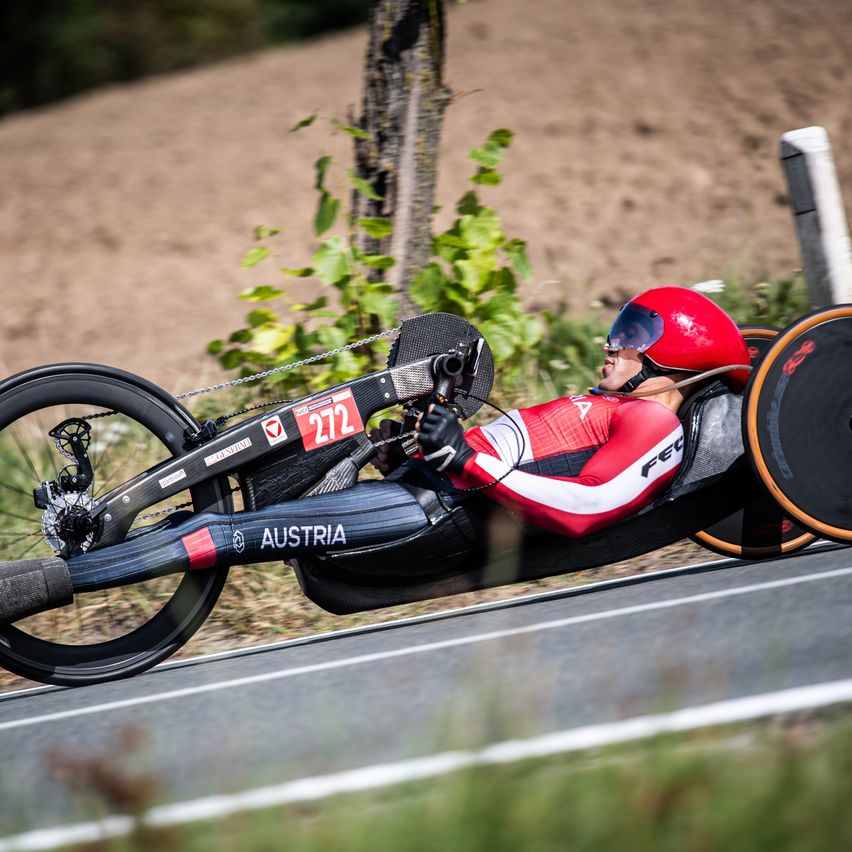Ein Athlet aus Österreich mit rotem Helm und rot-weißem Anzug fährt auf einer Straße mit einem Handrad. Das Fahrrad hat die Nummer 272 und das Wort Österreich darauf.