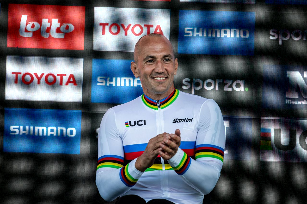 Ein Mann in einem Radtrikot lächelt und klatscht, während er vor einer Kulisse mit Logos steht.