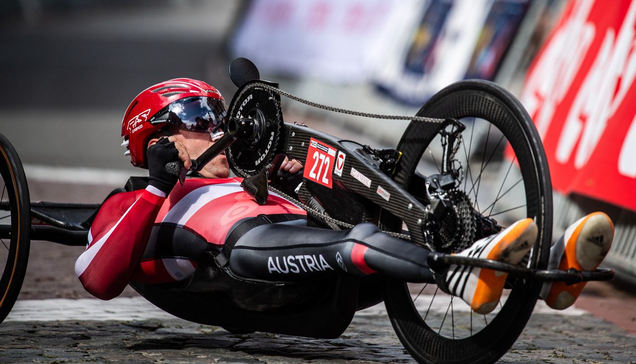 Ein Mann liegt auf einem Fahrrad mit der Nummer 272 auf dem Rücken und dem Wort Österreich auf der Hose. Er trägt einen roten Helm und Handschuhe.