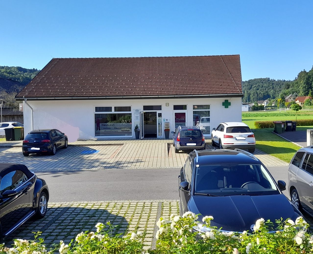 Ein weißes Gebäude mit braunem Dach hat vor dem Eingang geparkte Autos, mit einem grünen Kreuzzeichen darüber. Rechts befinden sich zwei Mülleimer.