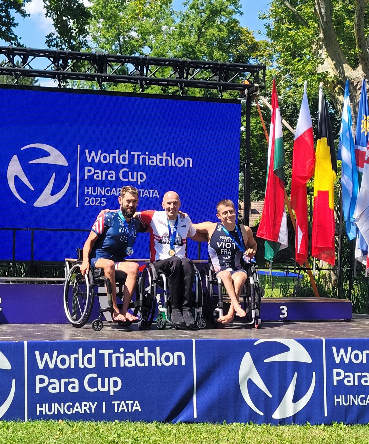 Drei Männer in Rollstühlen posieren auf einem Podium mit Medaillen. Sie sind beim World Triathlon Para Cup in Ungarn, 2025. Flaggen und ein großer blauer Banner hinter ihnen.