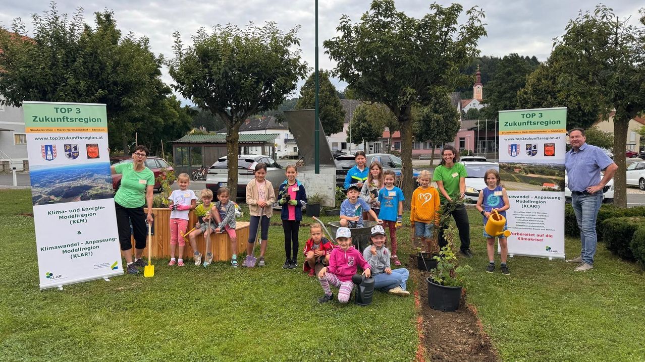 Eine Gruppe von Kindern und Erwachsenen pflanzt Bäume in einem Park. Sie tragen Handschuhe und halten Schaufeln. Ein Mann und eine Frau stehen hinter ihnen, einer hält eine Gießkanne. Der Park hat grünen Rasen, Bäume und geparkte Autos im Hintergrund.