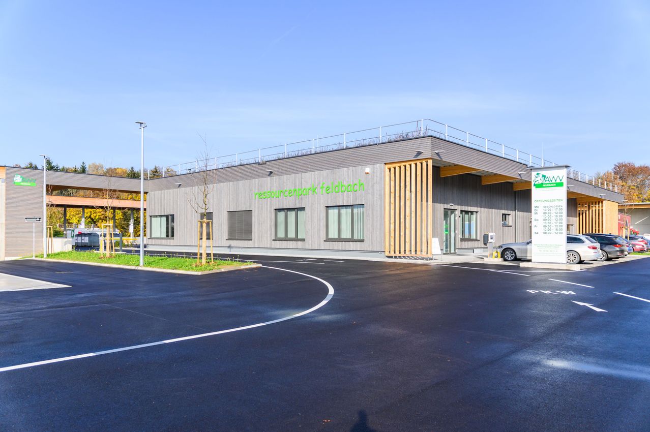Ein modernes Gebäude mit der Aufschrift 'Ressourcenpark Feldbach' an der Seite. Es hat eine Dachterrasse, Glasfenster und einen Parkplatz mit einem geparkten Auto.