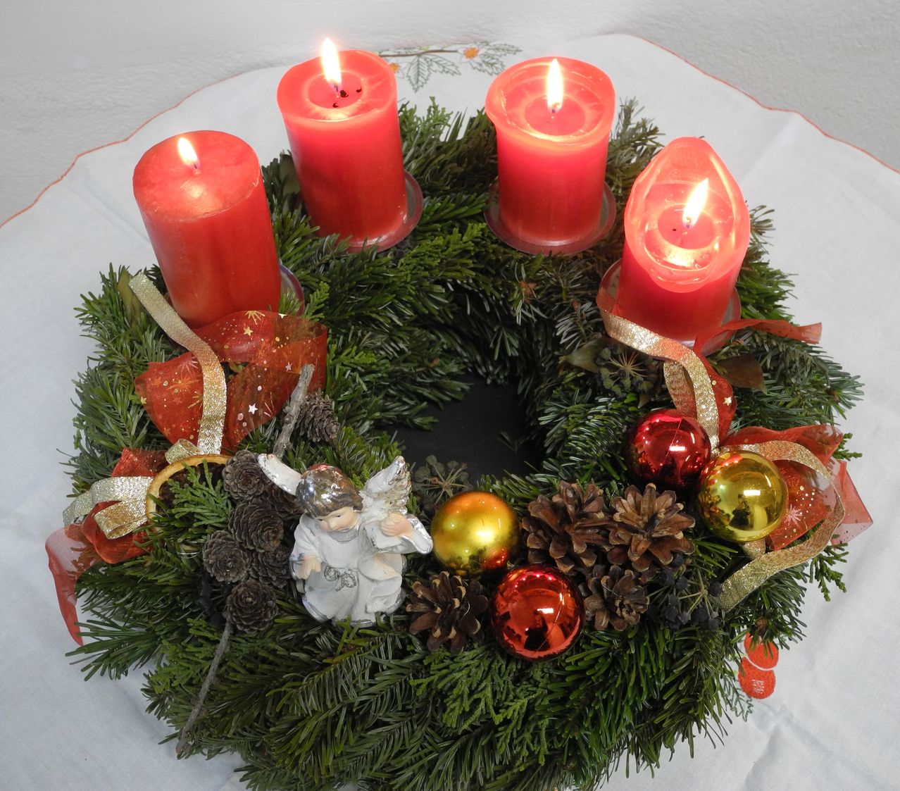 Ein Kranz mit vier roten Kerzen, einem Engel und verschiedenen Ornamenten wie Tannenzapfen, einer Zitrusscheibe und Weihnachtskugeln, auf einer weißen Tischdecke platziert.