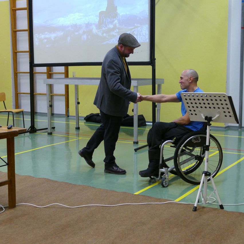 Zwei Männer, einer im Rollstuhl und der andere in formeller Kleidung, schütteln sich in einer Turnhalle die Hände. Dahinter zeigt eine Projektionsleinwand eine Berglandschaft.