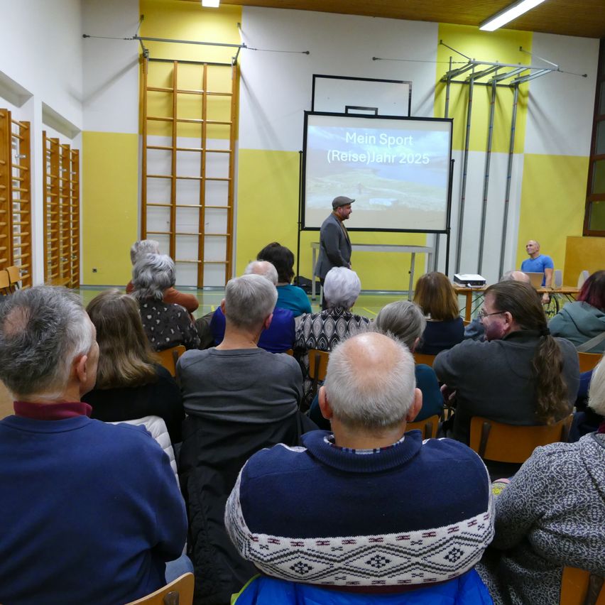Ein Saal mit gelben und weißen Wänden. Ein Mann steht vor einer Projektionsleinwand. Er trägt einen Hut und eine Jacke. Die Leinwand zeigt 'Mein Sport (Reise) Jahr 2025'. Eine Leiter ist an der Wand. Eine Gruppe von Menschen sitzt auf Stühlen.