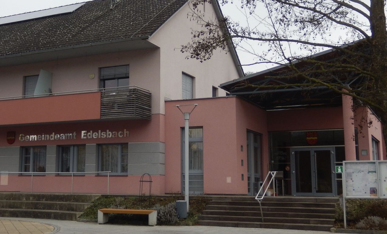 Die Vorderseite eines rosa Gebäudes mit dem Wort Edelsbach darauf. Eine Bank und eine Laterne befinden sich auf der linken Seite. Eine Treppe führt zum Eingang.