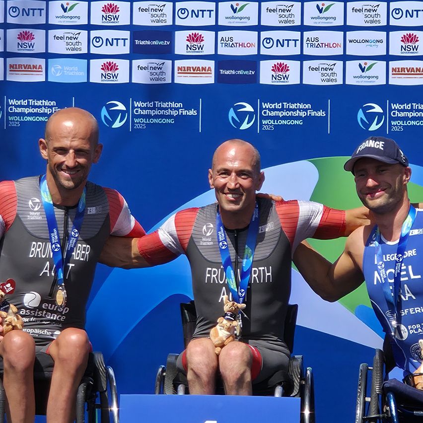 Drei Männer, alle mit Medaillen, sitzen auf einem Podium bei den World Triathlon Championship Finals in Wollongong, Australien. Der Mann links trägt ein Trikot mit der Aufschrift 'Brun', das Trikot des Mannes in der Mitte lautet 'Fruit' und das Trikot des Mannes rechts hat die Aufschrift 'Nicole'. Sie alle haben Lächeln auf den Gesichtern.