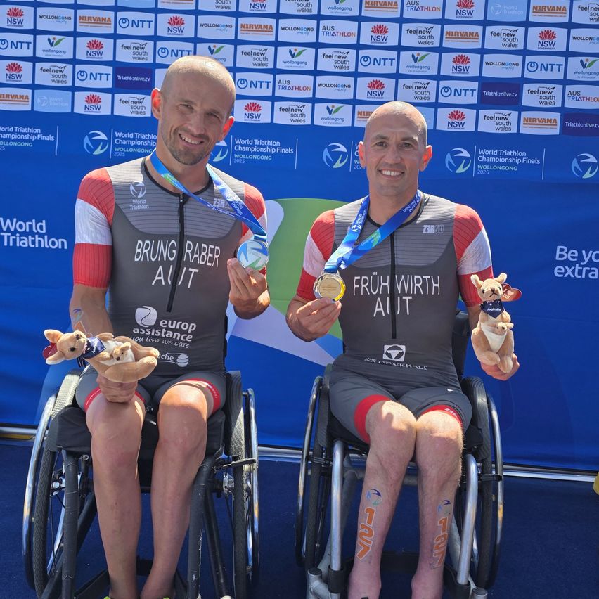 Zwei Männer im Rollstuhl halten ihre Medaillen und posieren lächelnd für ein Foto vor einem Banner. Der Mann auf der linken Seite trägt ein Brung Raber Australasia-Trikot und der Mann auf der rechten Seite trägt ein Fruh Wirth-Trikot. Beide haben Tattoos an den Beinen.
