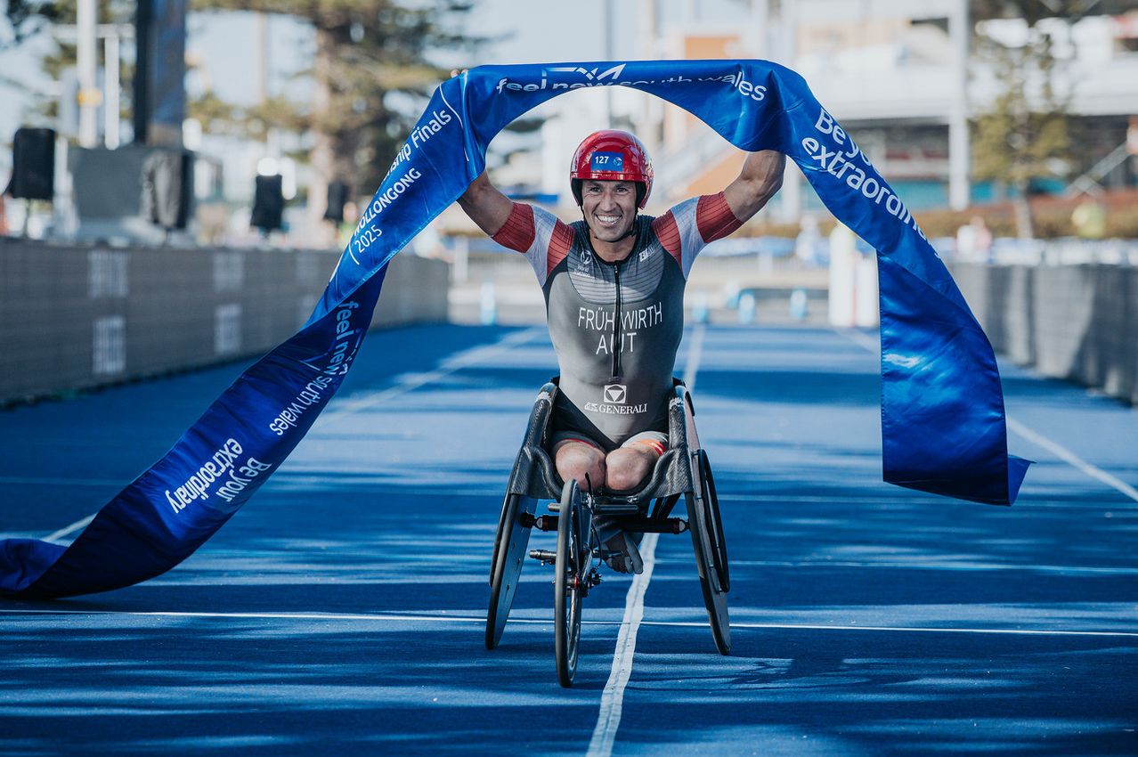 Ein Mann im Rollstuhl, mit rotem Helm, feiert mit einer blauen Schleife auf einer Rennstrecke.