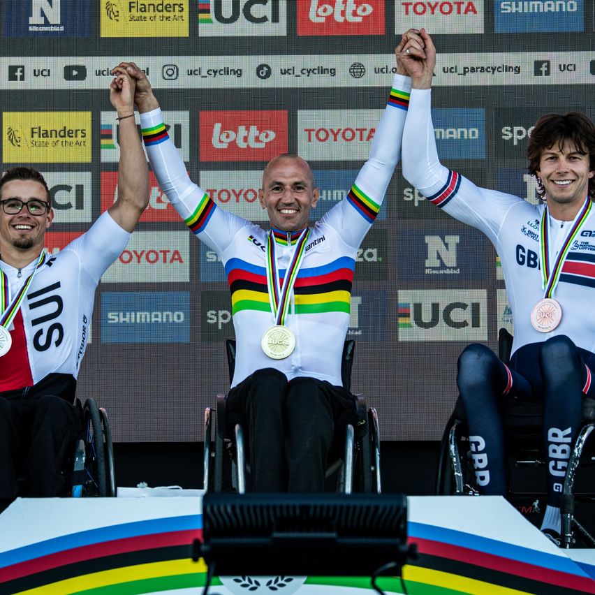 Drei Männer im Rollstuhl, die Radsportuniformen und Medaillen tragen, feiern auf einem Podest. Sie lächeln und heben die Hände. Hinter ihnen ist eine Wand mit verschiedenen Logos.