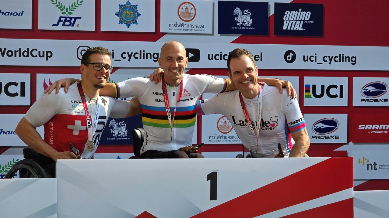 Three cyclists on the podium, two in white uniforms and one in a wheelchair, smiling for the camera with medals around their necks. Behind them, a wall displays various logos and sponsors.