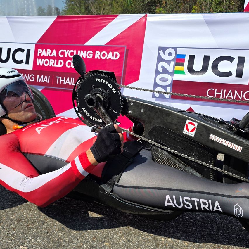 A man wearing a red and white suit lies on a recumbent bike on the road. The bike has the word Austria on it. He is holding a chain.