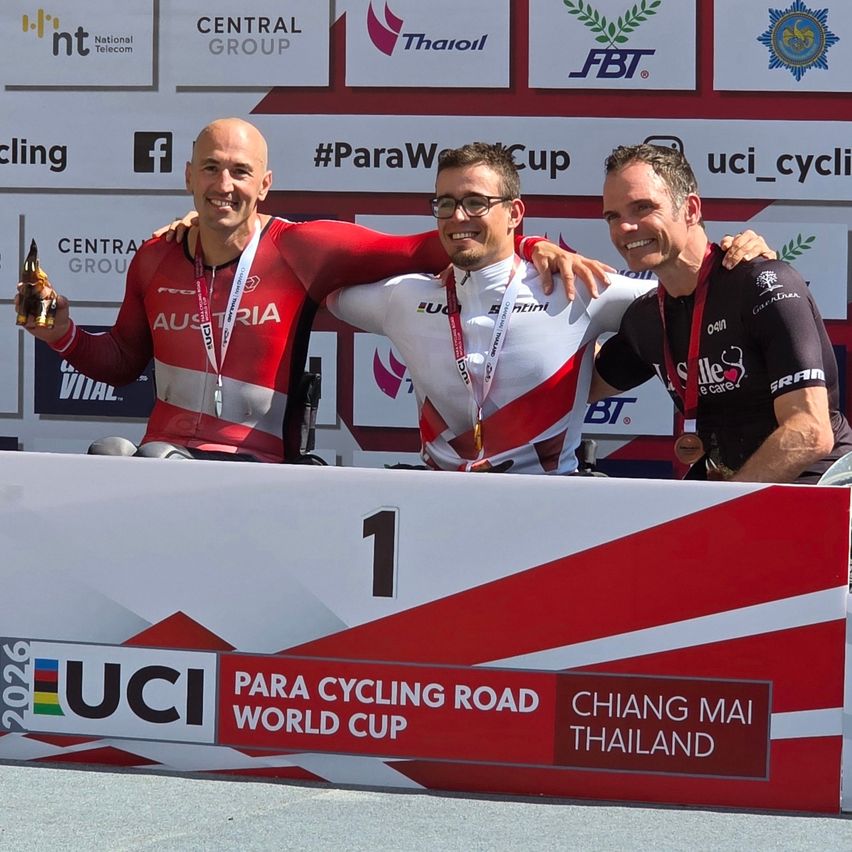 Three men celebrate on a podium at the UCI Para Cycling Road World Cup in Chiang Mai, Thailand, with one holding a trophy.