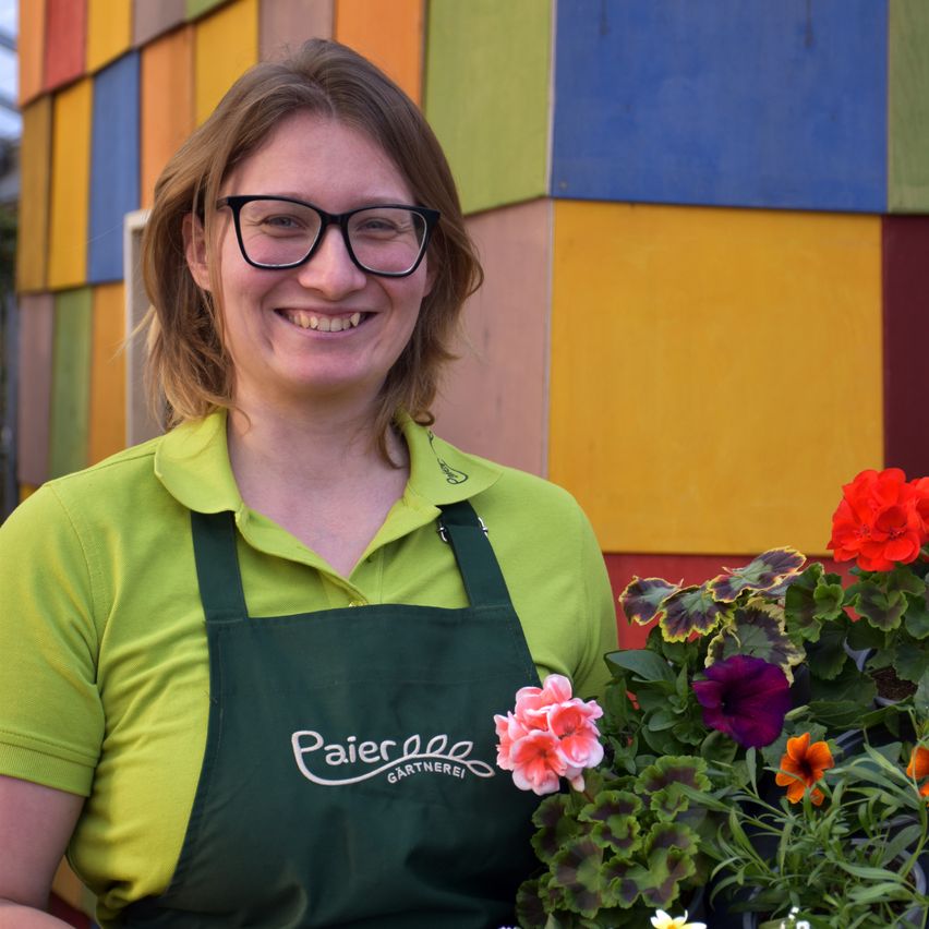 Eine Frau mit Brille und einer grünen Schürze mit weißen Buchstaben lächelt und hält eine Topfpflanze mit rosa, lila und orangefarbenen Blumen. Hinter ihr befindet sich eine farbenfrohe Wand, die aus quadratischen Paneelen in verschiedenen Farben besteht.