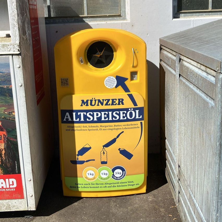 A yellow container for old cooking oil is placed outside a building. It has a sticker that reads 'MUNZER ALTSPEISEÖL' and icons indicating weight.