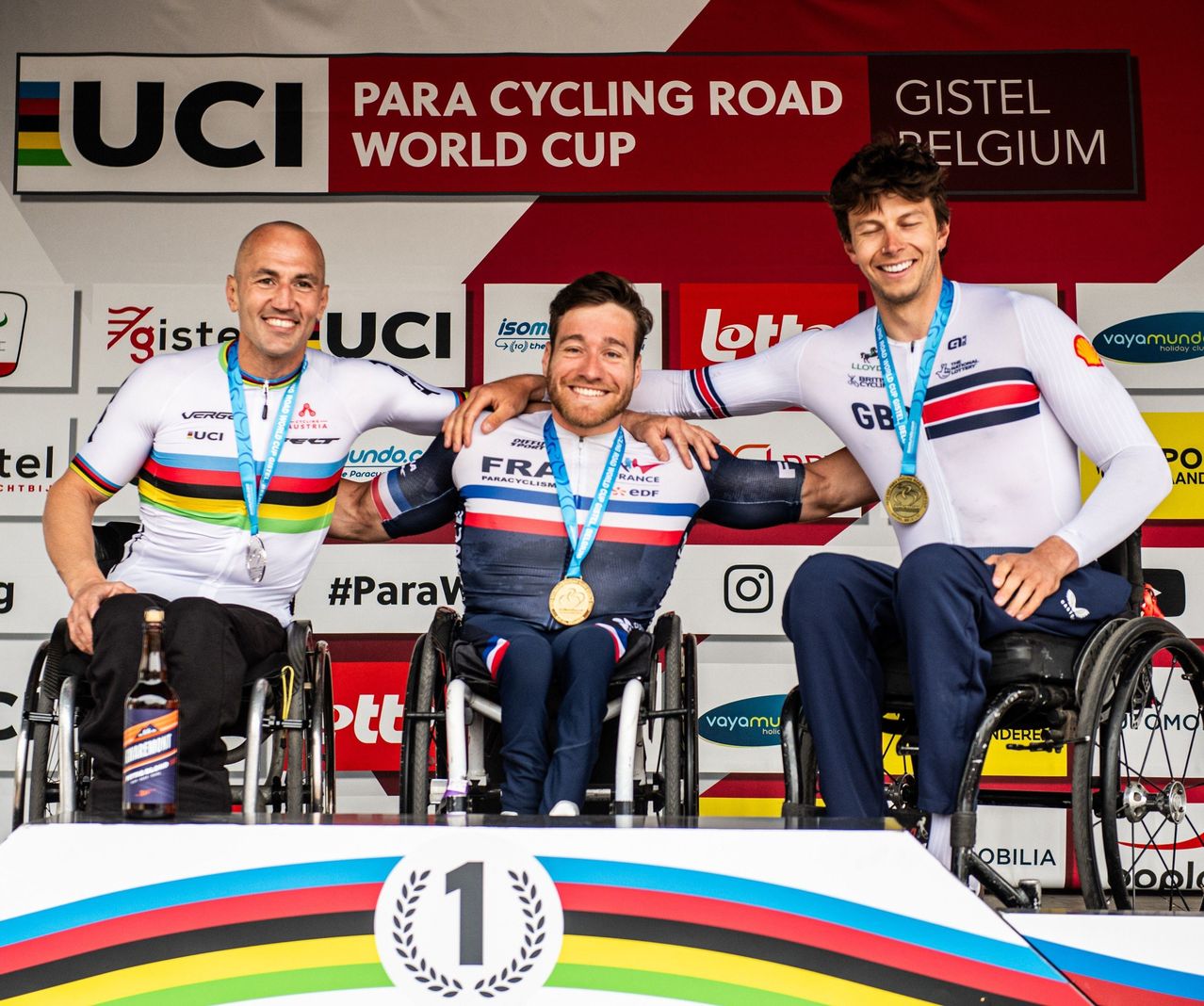 Three cyclists sit on a podium wearing cycling uniforms and medals. Behind them, a sign reads UCI PARA CYCLING ROAD WORLD CUP, indicating a cycling competition.