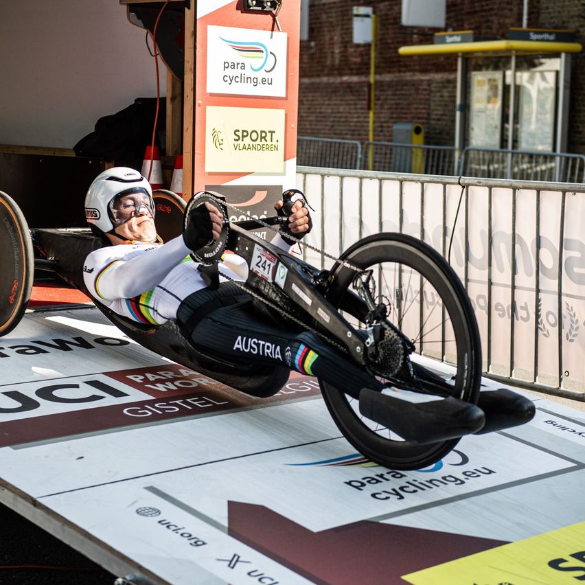 An athlete from Austria, wearing a white helmet and cycling gear, lies on a recumbent bicycle, preparing for a race on a track marked 'paracycling.eu'.