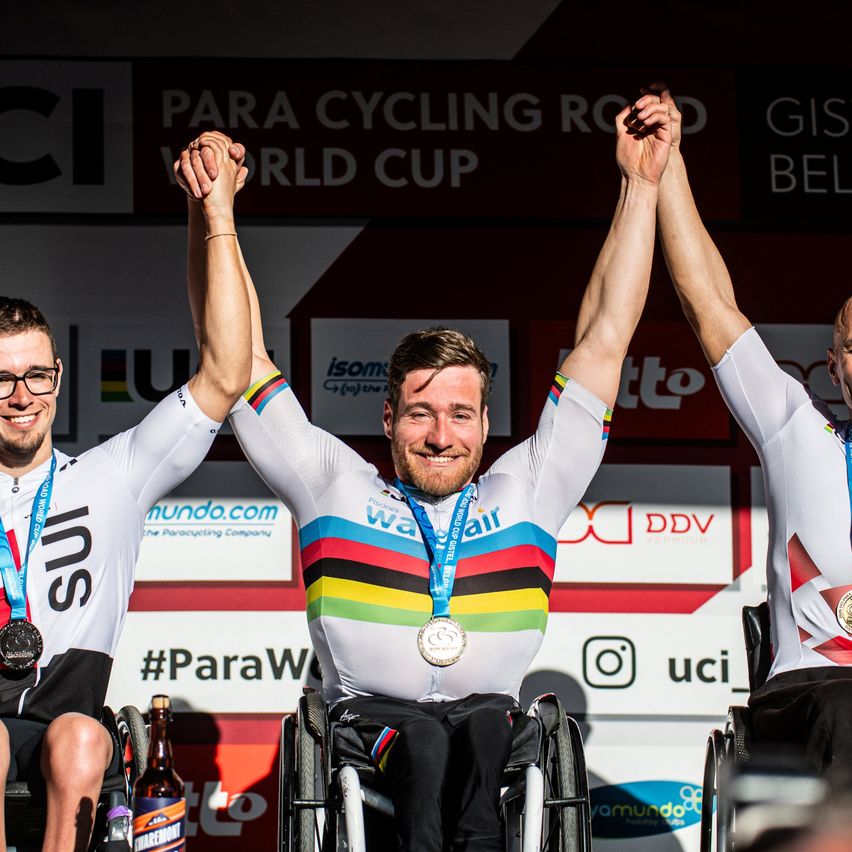 Three men in wheelchairs celebrate with medals and smiles on a podium. The man in the center wears a rainbow jersey and a gold medal. Behind them, banners display 'PARA CYCLING WORLD CUP' and 'GIS'.