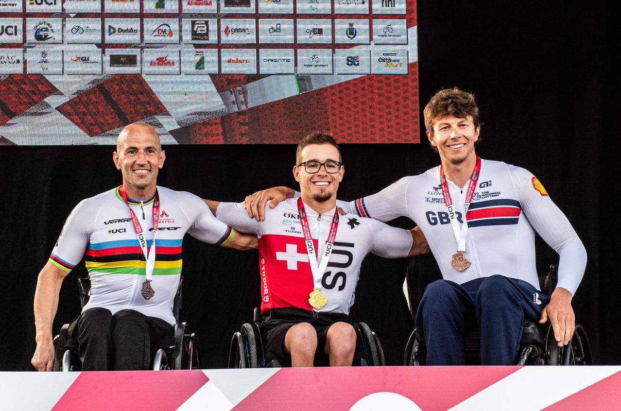 Three men in wheelchairs pose for a photo with medals on a stage. They are smiling and wearing cycling jerseys. Behind them is a screen with logos.
