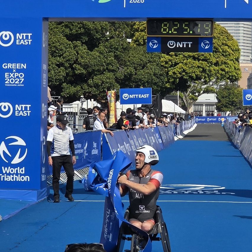 An athlete in a wheelchair finishes a triathlon, surrounded by spectators, with a large digital clock showing the time above.