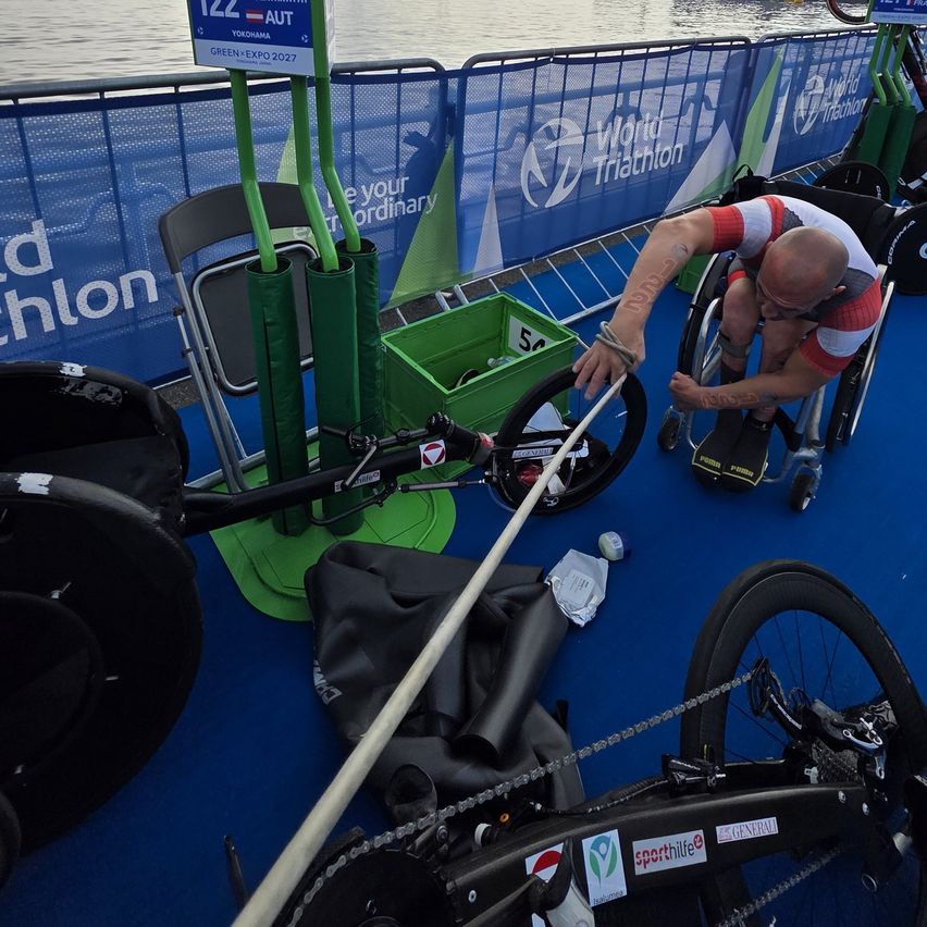 A man in a wheelchair works on his bike in a triathlon event with a water background and green barriers.