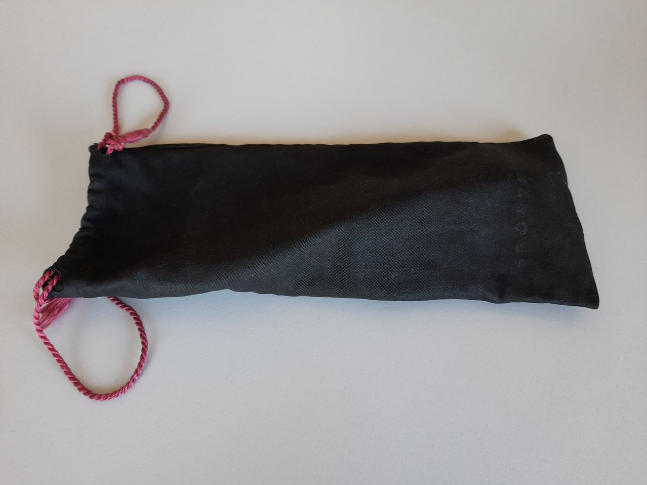Ein schwarzer Stoffbeutel mit einem roten Zugband liegt auf einer weißen Oberfläche. Der Beutel ist geschlossen und scheint eine glatte Textur zu haben.