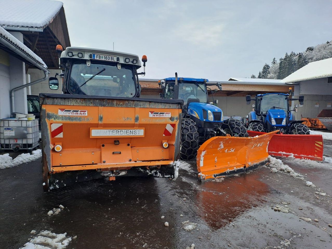 Ein orangefarbener Schneepflug steht vor einem Gebäude, mit anderen Traktoren in der Nähe. Der Schneepflug hat den Schriftzug Hydrac und ist als Winterdienst gekennzeichnet. Das Gebiet ist mit Schnee bedeckt.