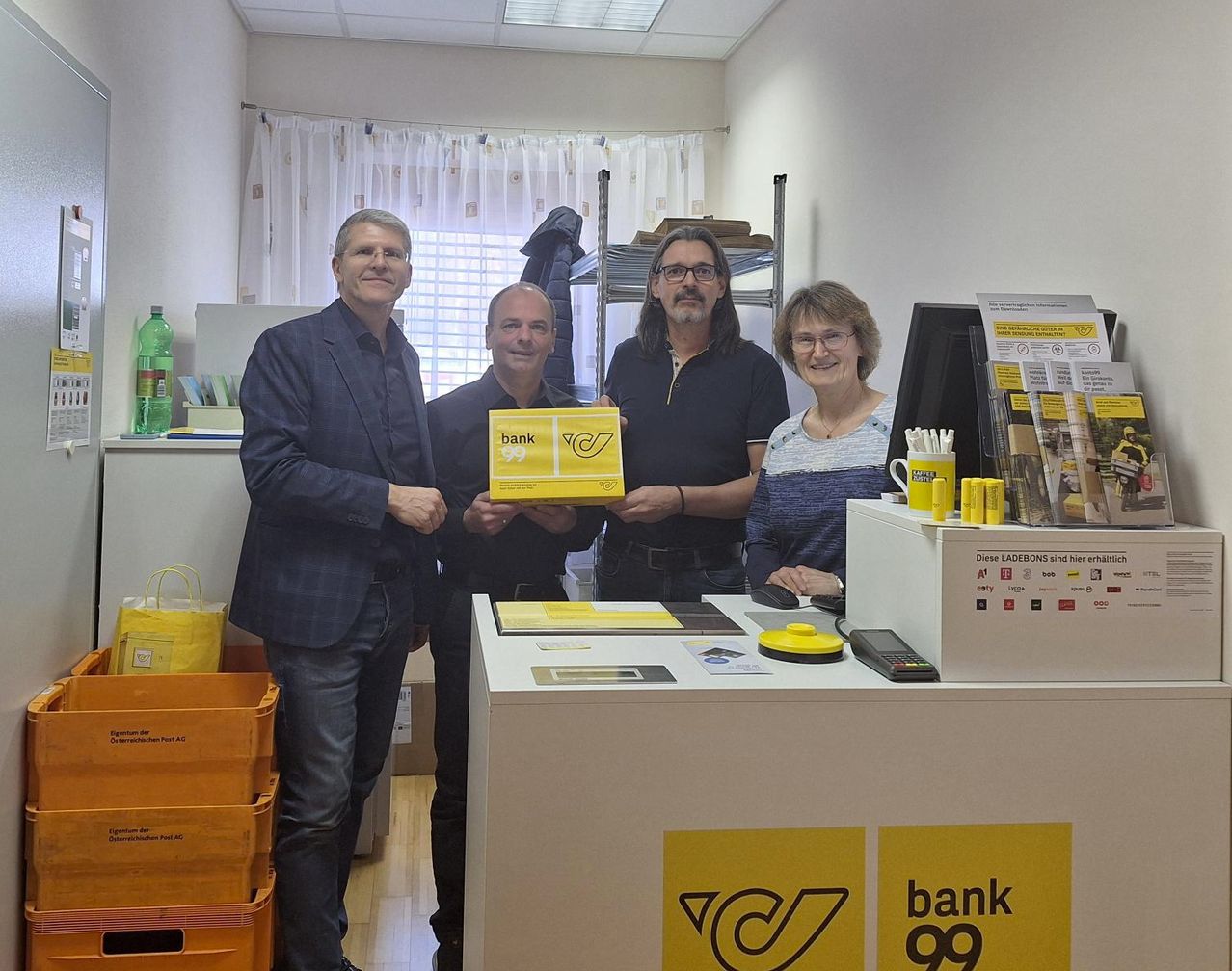 Vier Personen stehen in einem Bankbüro und lächeln für ein Foto. Eine gelbe Box wird von zwei Männern in der Mitte gehalten. Hinter ihnen arbeitet eine Frau an einem Computer.