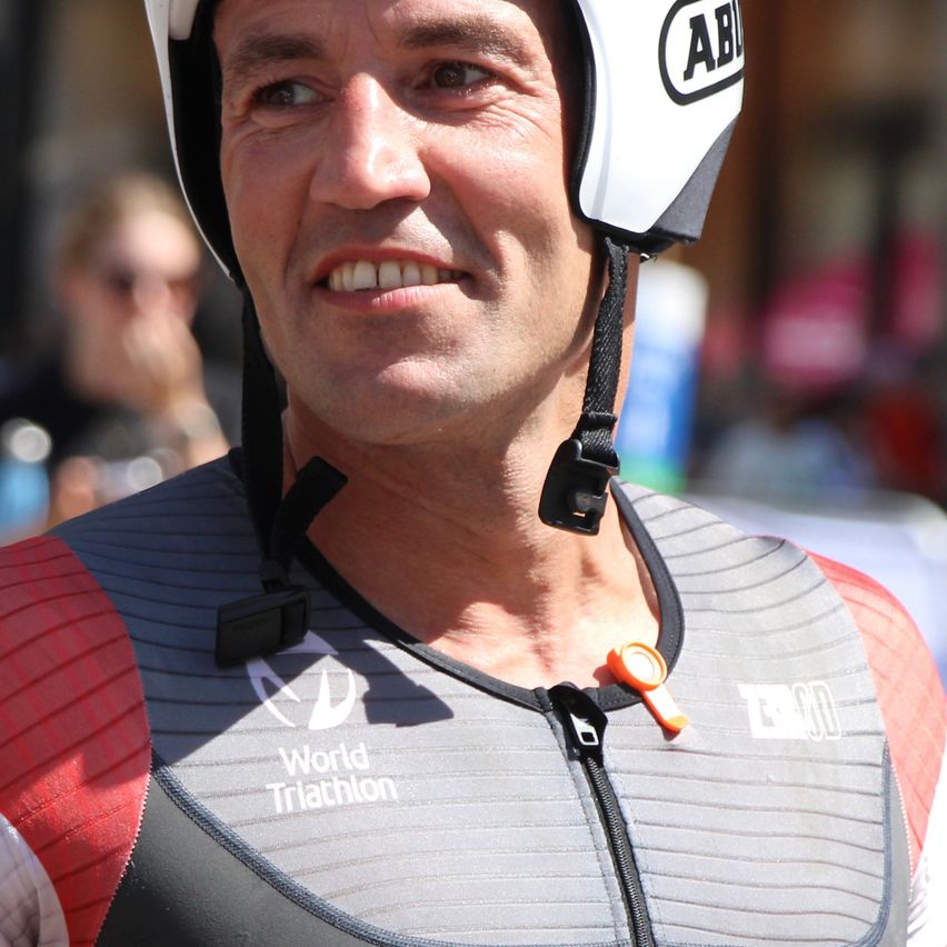 Ein Mann mit einem weißen Helm mit den Buchstaben AB und einem roten und grauen Triathlon-Trikot lächelt in die Kamera.