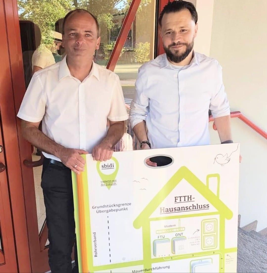 Two men stand in front of a building, holding a sign with a house and the words 'FTTH-Hausanschluss'. One man has a beard, and both are smiling.