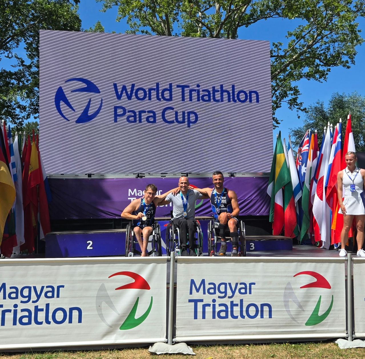 Drei Athleten in Rollstühlen sitzen auf einer Bühne mit Flaggen dahinter, wahrscheinlich bei einem Triathlon-Event.